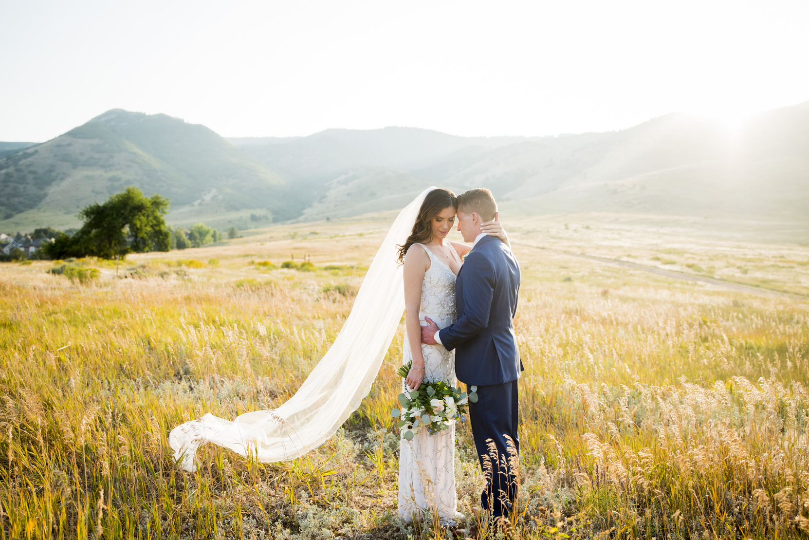 The bride and groom are standing in a field of tall grass with the sun shining down on them and the mountains in the background.