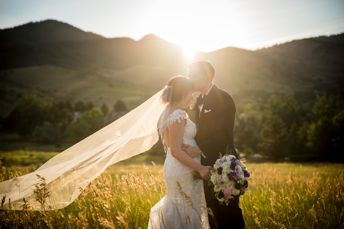 A groom kisses his bride on the forehead with the sun setting behind them in a field.