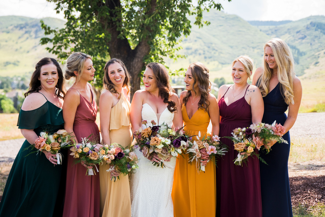 A bride and her bridesmaids in colorful dresses under a tree.