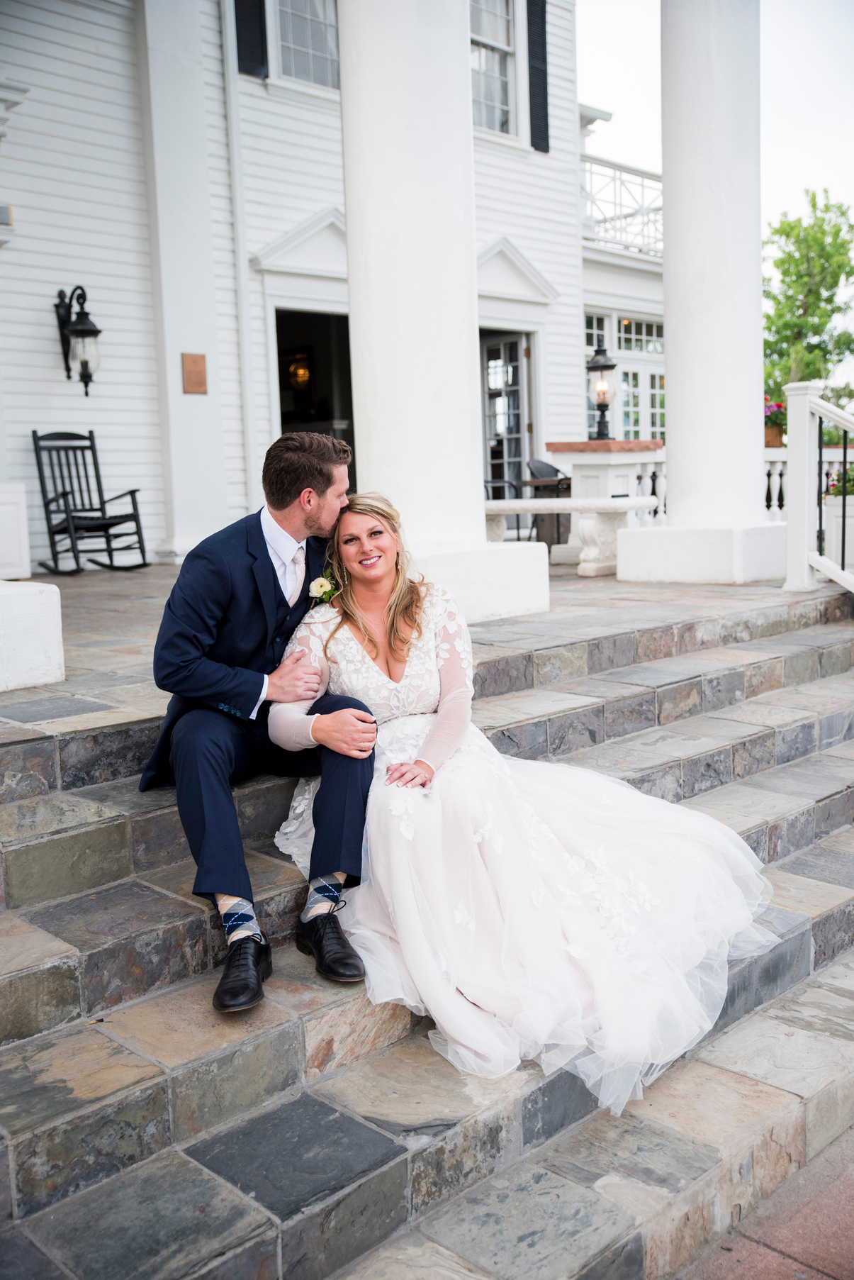 A bride and groom sitting on steps in front of white house at The Manor House in Littleton, Colorado.
