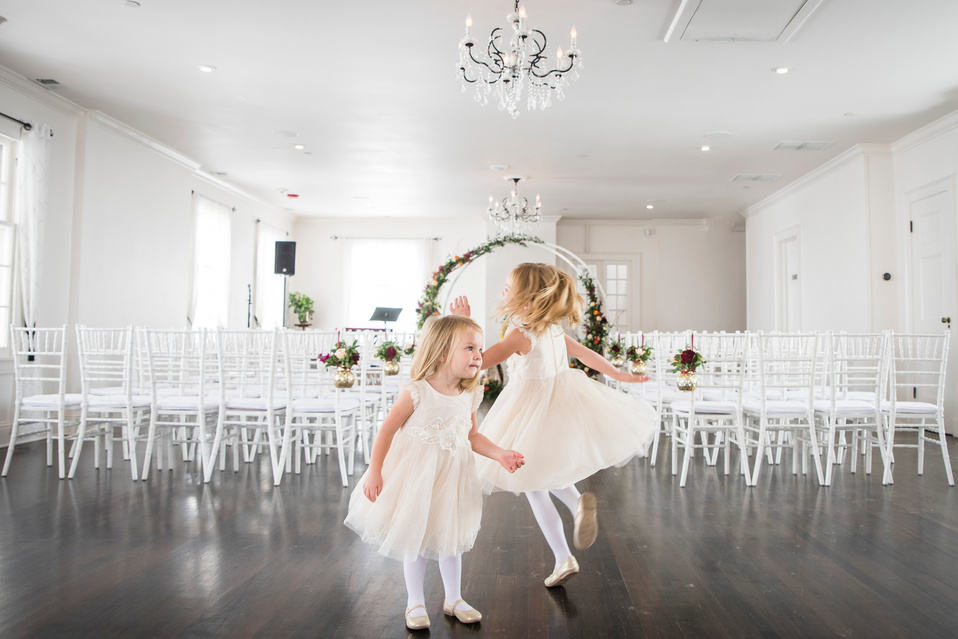 Two little flower girls in white dresses dancing in the middle of an empty room.