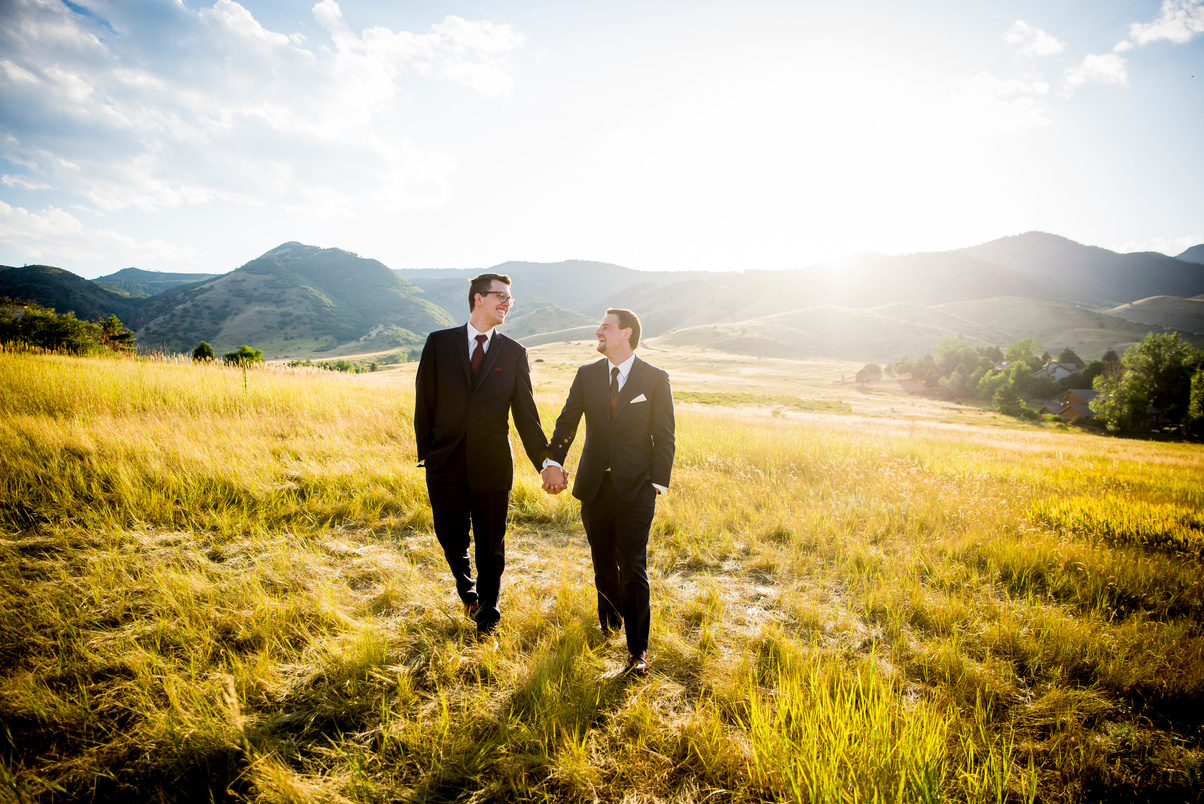 Two grooms in suits holding hands in the middle of a field.
