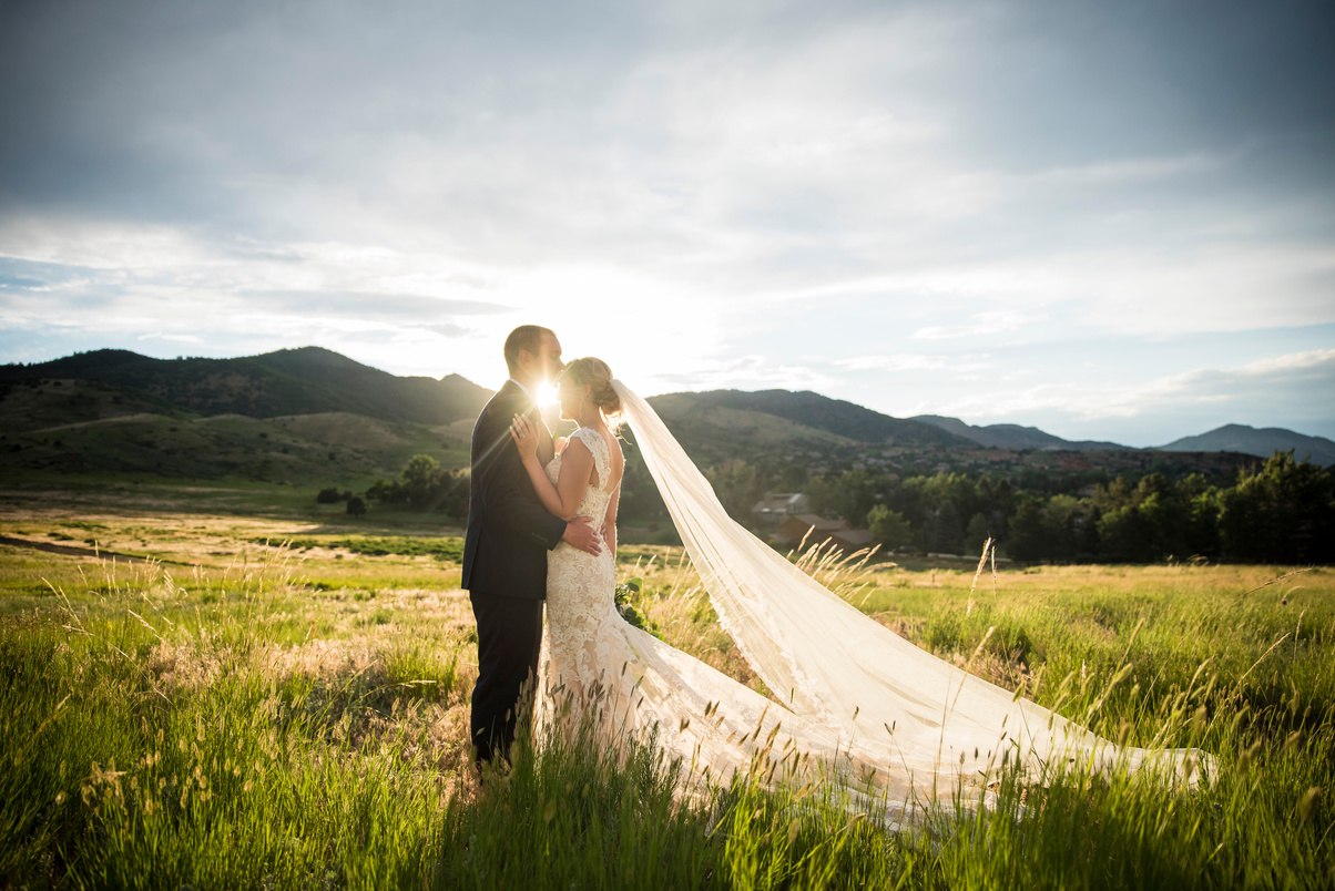 The bride and groom are standing in the middle of a grassy field with the sun setting behind them.
